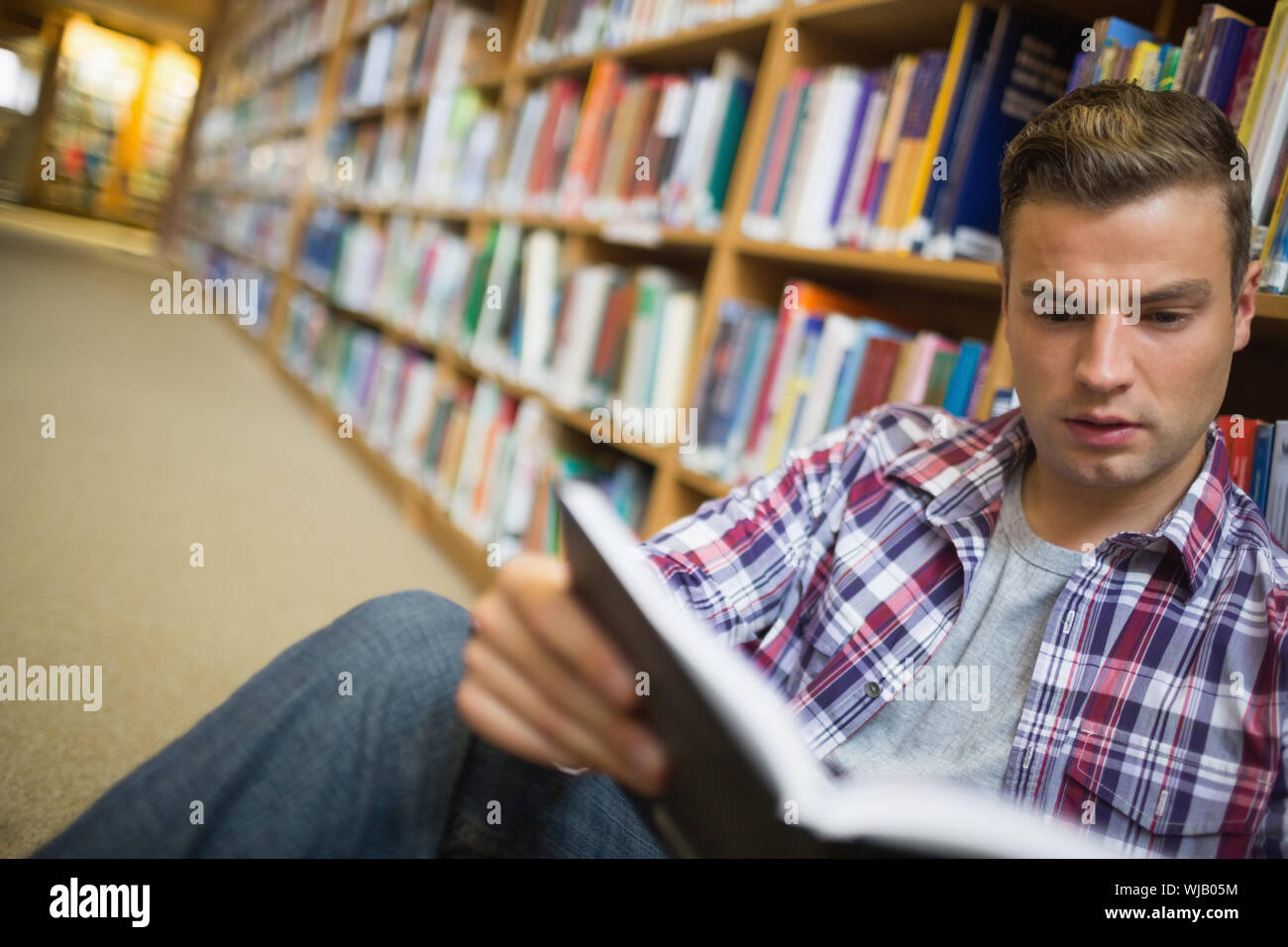 Serious young student sitting on library floor reading book Stock Photo