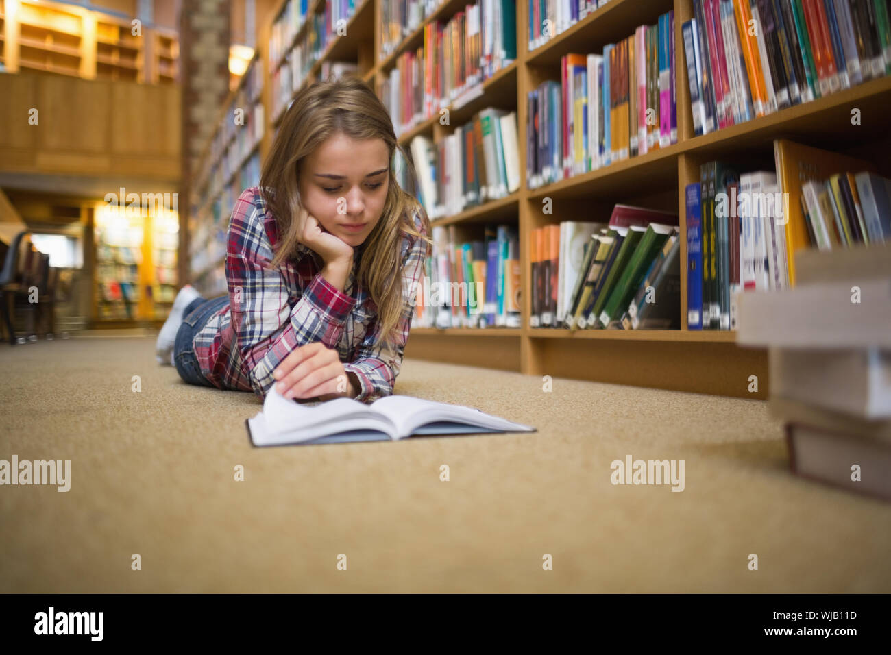Pretty focused student lying on library floor reading book Stock Photo