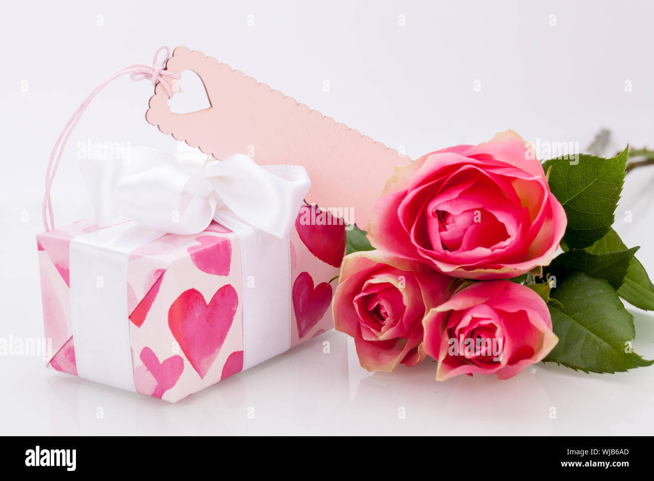 Close-up of a gift box wrapped in decorative paper with pattern with heart shapes, with an empty tag, next to three roses, symbol of love and apprecia Stock Photo