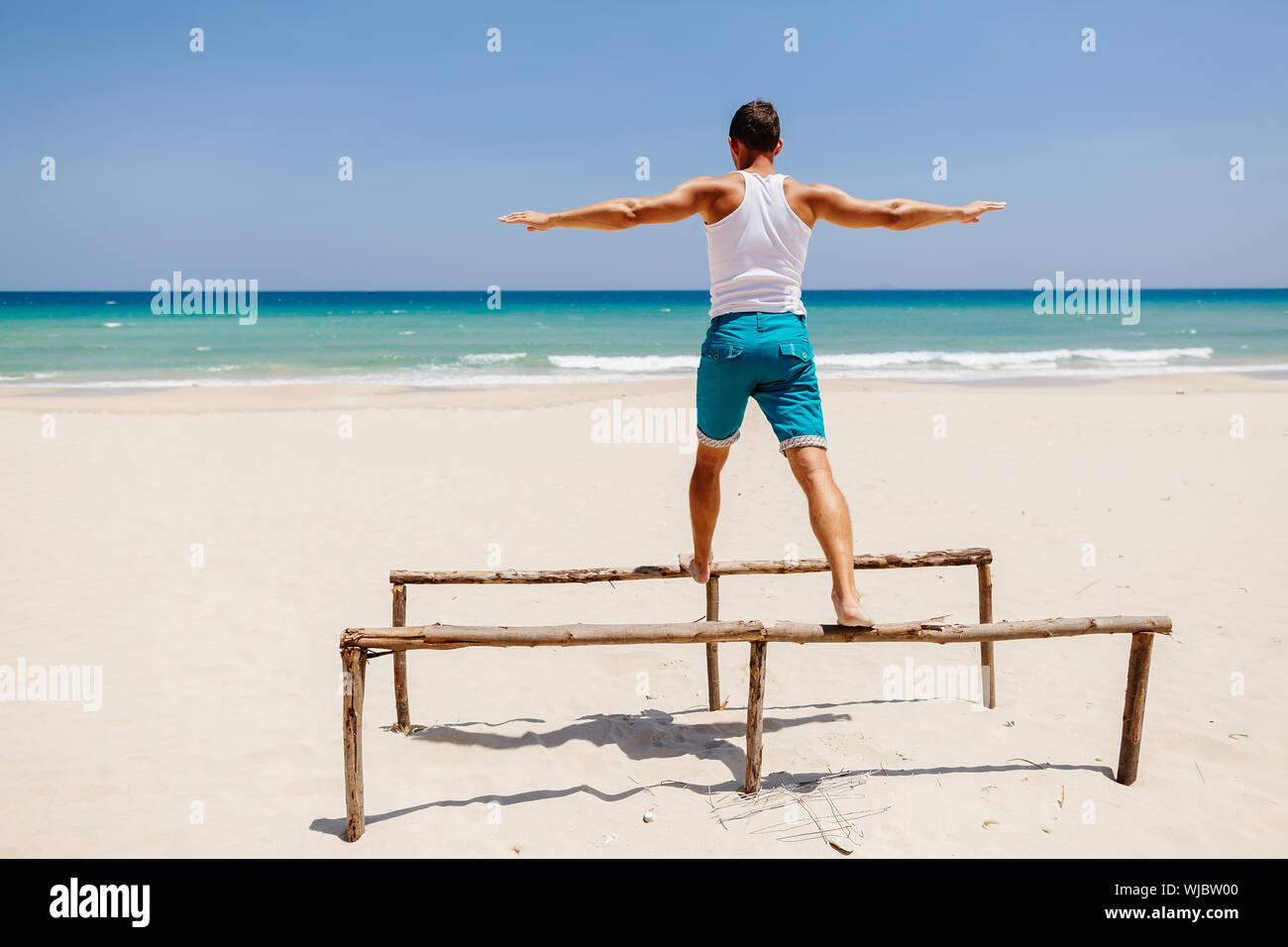 Man With Arms Outstretched Balancing On Wooden Structure At Beach During Sunny Day Stock Photo