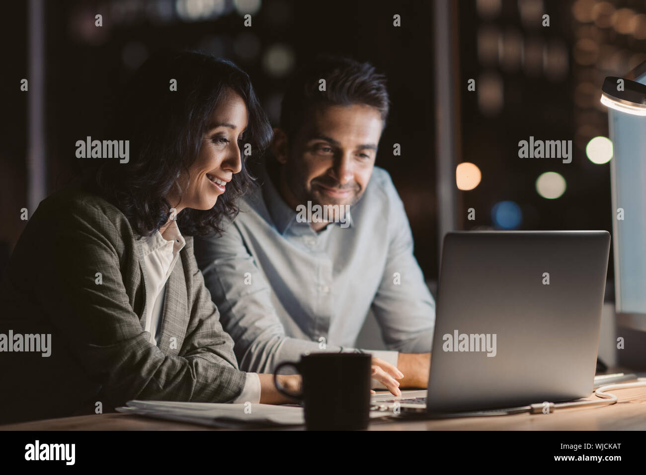 Two smiling office colleagues using a laptop while working overtime Stock Photo