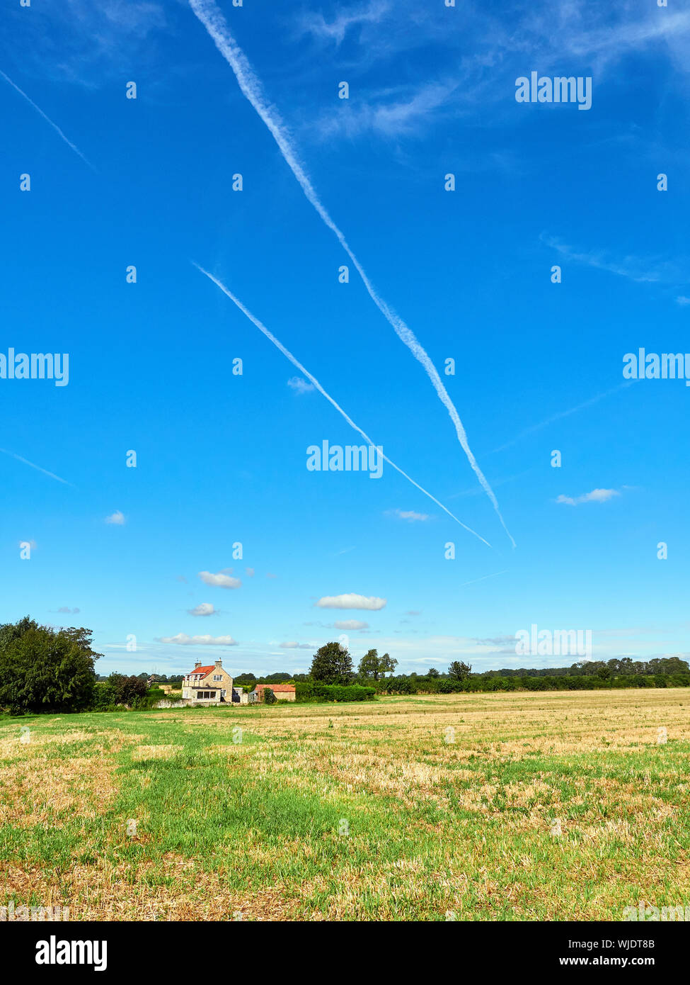 Aeroplane contrails in a blue sky over a stubble field with a stone house in the distance Stock Photo