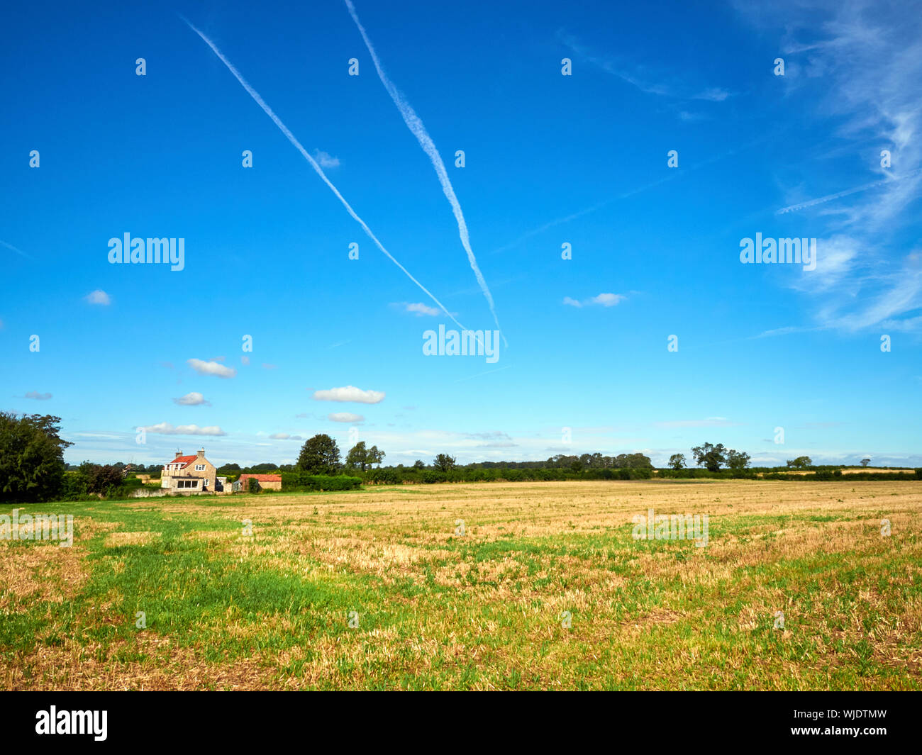 Aeroplane contrails in a blue sky over a stubble field with a stone house in the distance Stock Photo