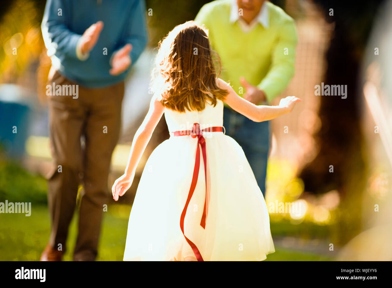 Young girl in a party dress runs towards two men in a garden. Stock Photo