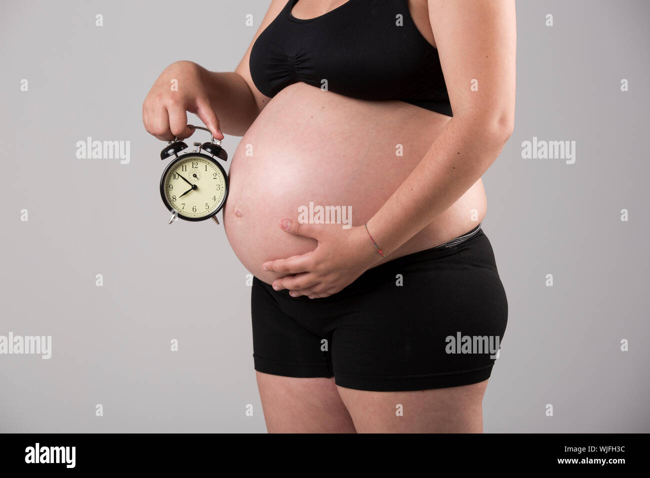 Pregnant woman showing her belly with a clock, over a gray background Stock Photo