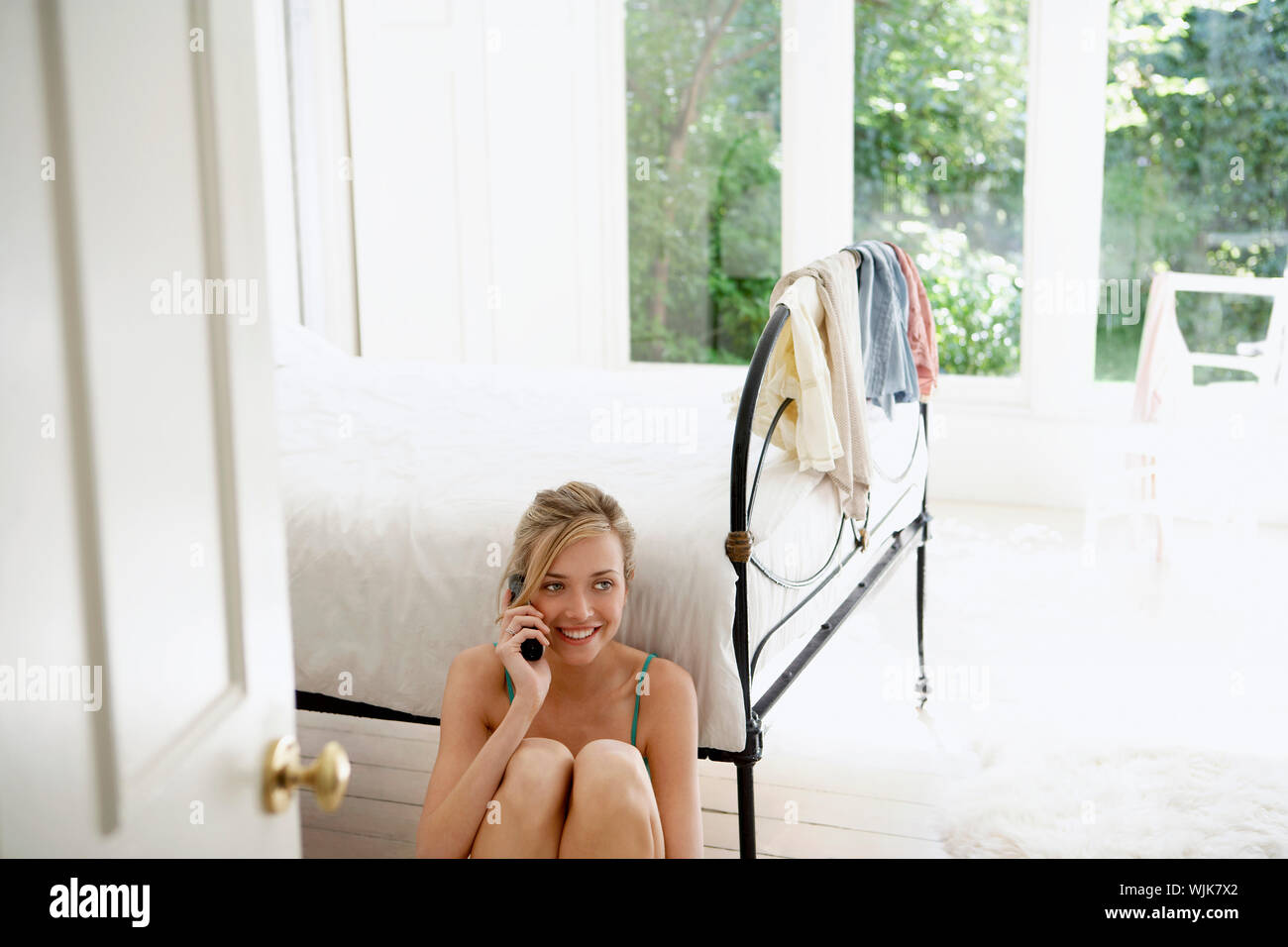 Happy young woman using mobile phone while sitting by bed Stock Photo
