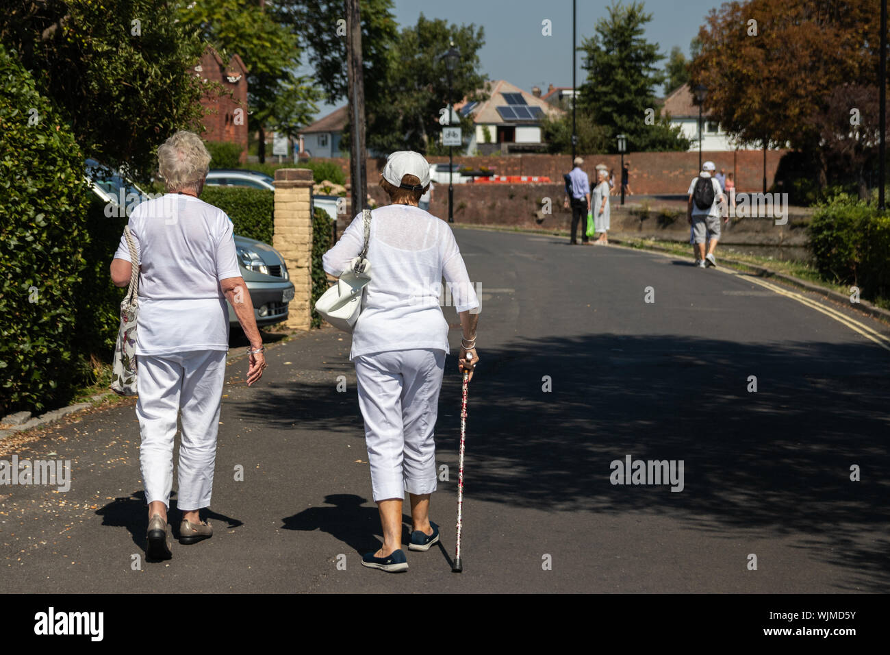 Two elderly women walking down a street, one of the women is using a walking stick or walking aid Stock Photo