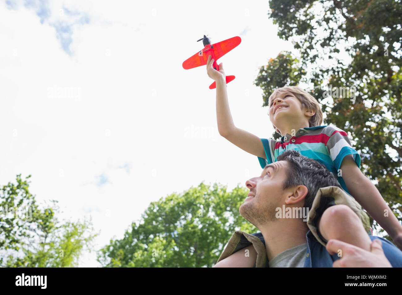 Low angle view of a boy with toy aeroplane sitting on father's shoulders at the park Stock Photo
