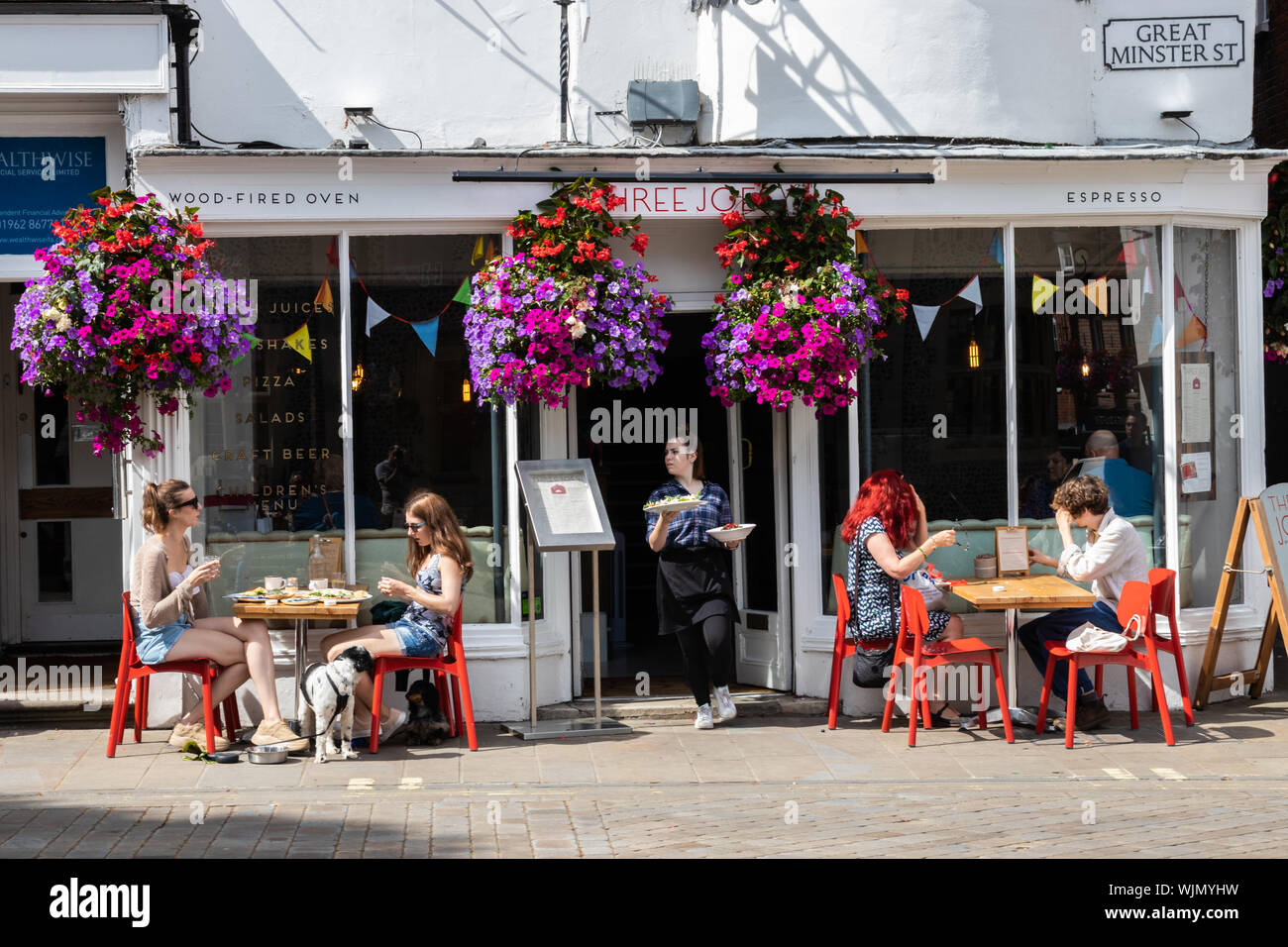 Winchester, Hampshire, UK the front of a cafe with people sitting at tables in the sun enjoying food and a coffee with a waitress carrying Stock Photo