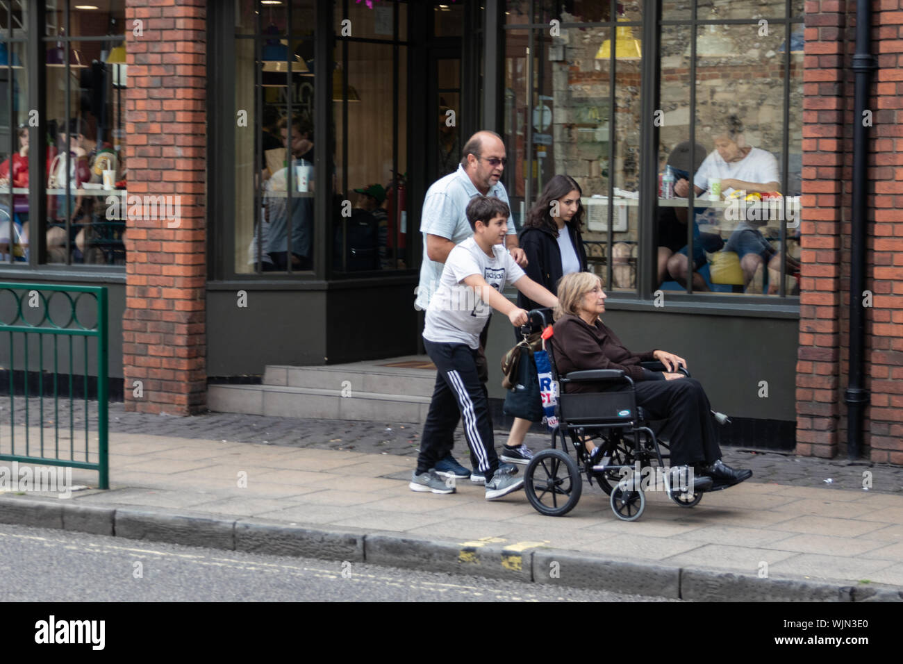 A young boy pushing an elderly lady in a wheelchair in a street past shop windows Stock Photo