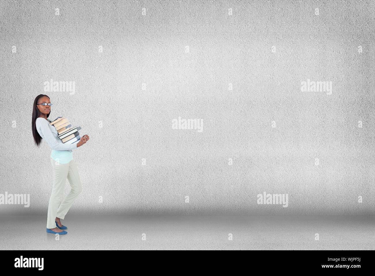 Side view of young woman carrying a pile of books against grey room ...