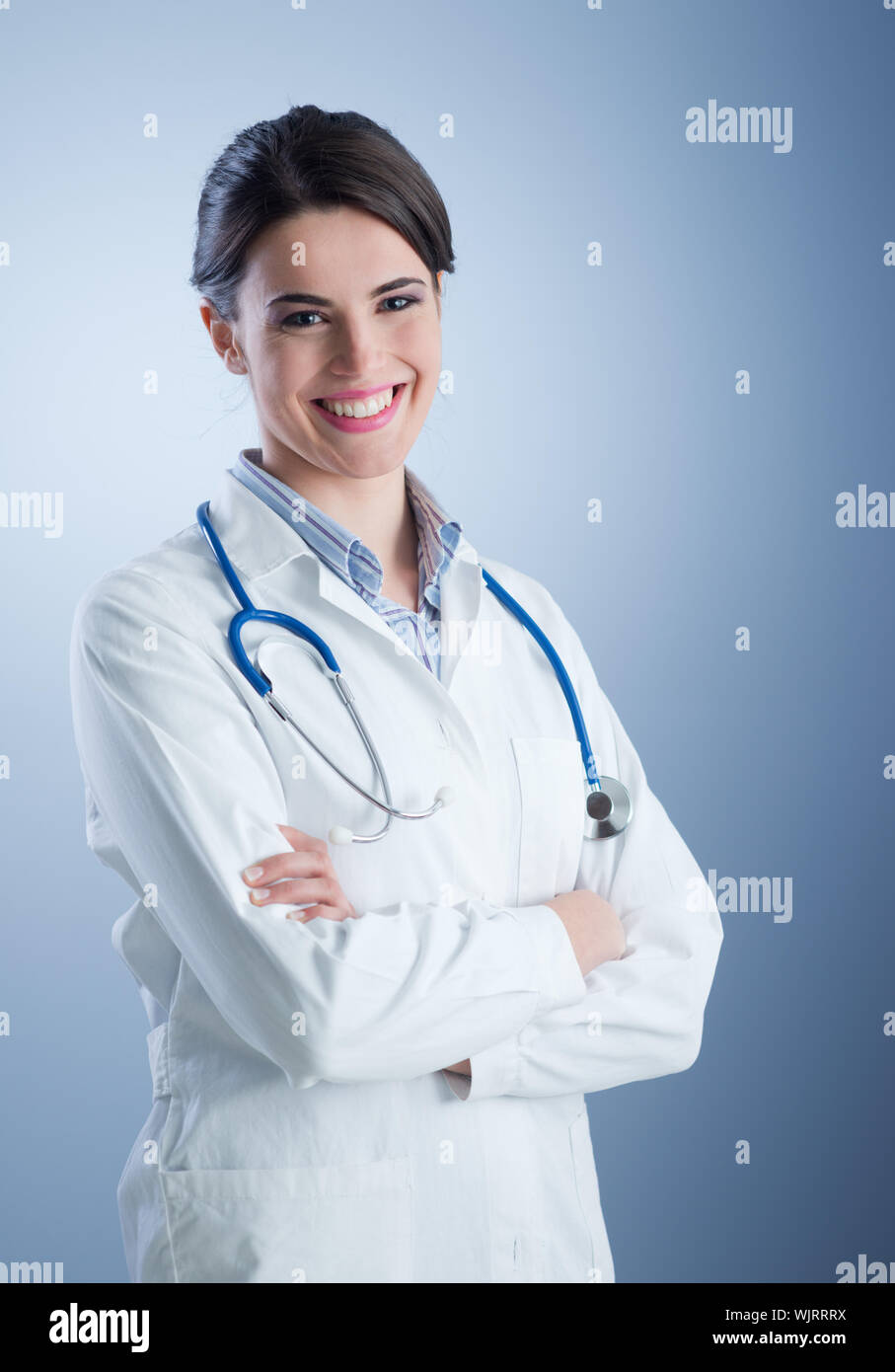 Self confident professional doctor stands with arms crossed, wears white  medical gown with stethoscope, thinks about work positively, poses against  big window. Healthcare and occupation concept - a Royalty Free Stock Photo