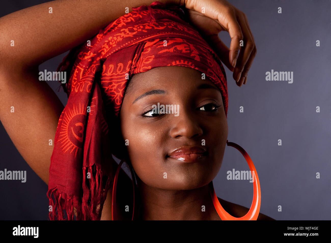 Beautiful African-American woman wearing a traditional tribal red orange head scarf and big orange hoop earrings, with arm over her head, isolated. Stock Photo
