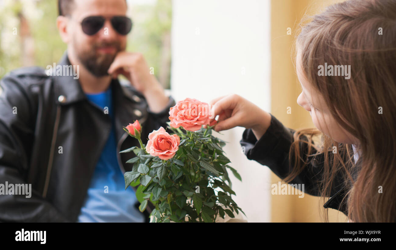 Girl touches the roses that stand on the table of the restaurant’s summer terrace. In the background, in the unfocusing of her, Dad looks and smiles Stock Photo