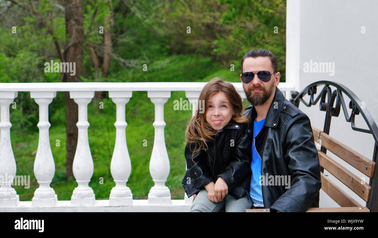 Fashionable stylish family portrait. Daughter sits on her father's lap and shows her tongue. Time of mischief. Time together. Family look. Urban casua Stock Photo