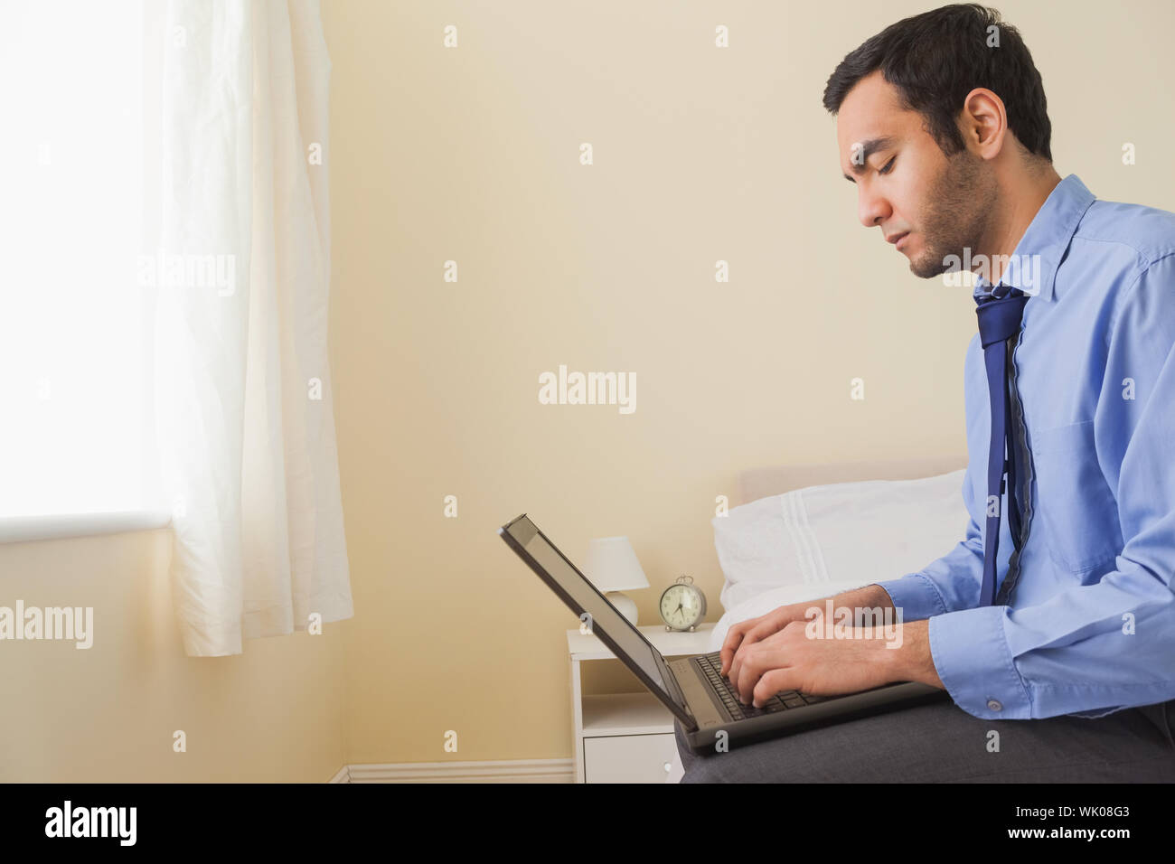 Unsmiling man using a laptop sitting on a bed Stock Photo