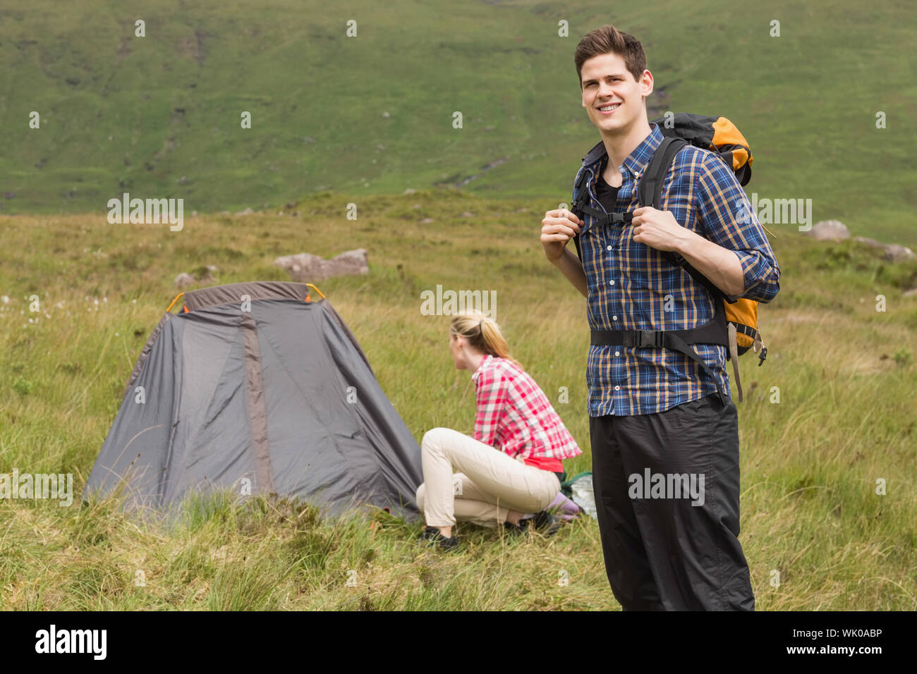 Smiling man carrying backpack while girlfriend is pitching tent Stock Photo