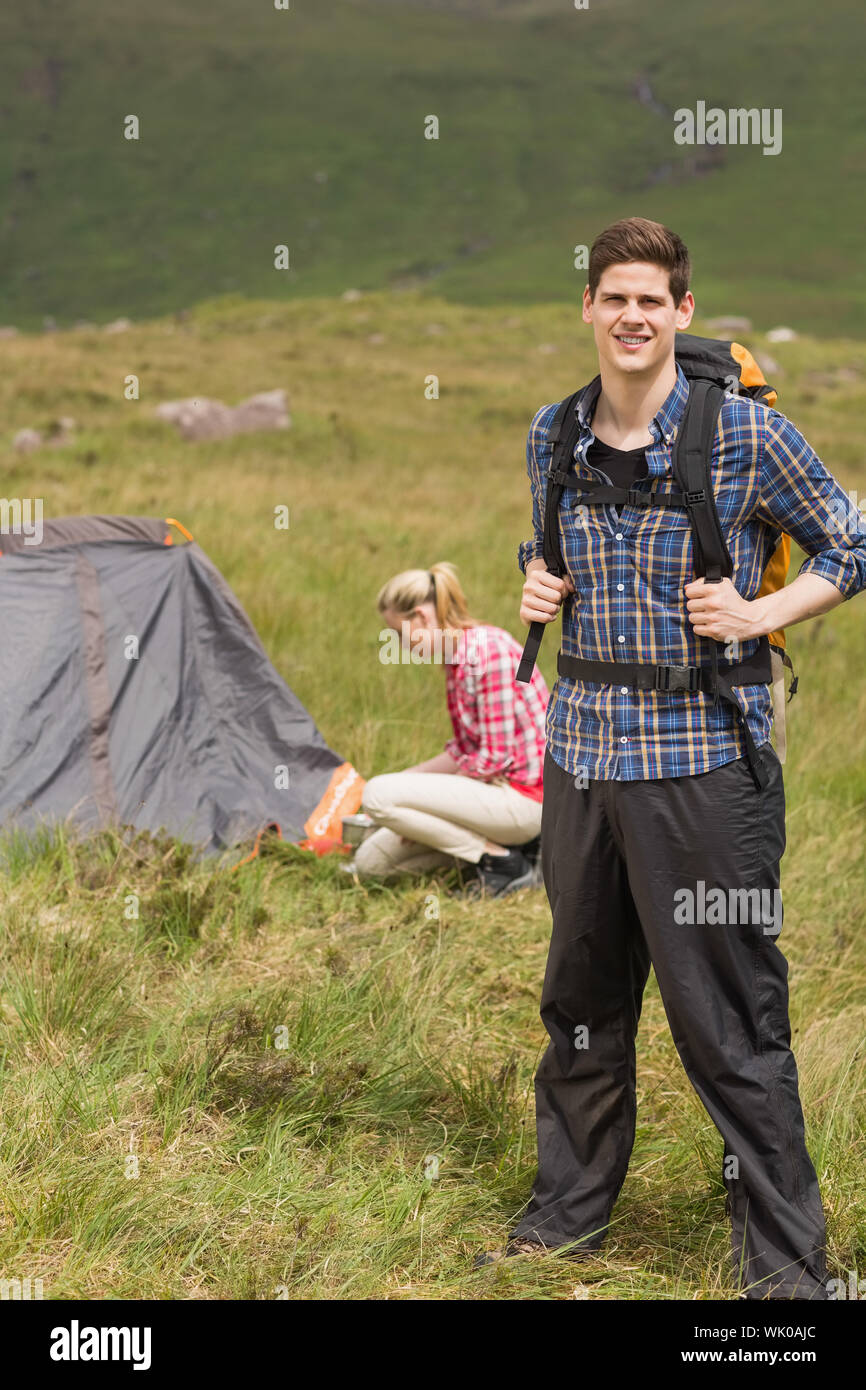 Happy man carrying backpack while girlfriend is pitching tent Stock Photo