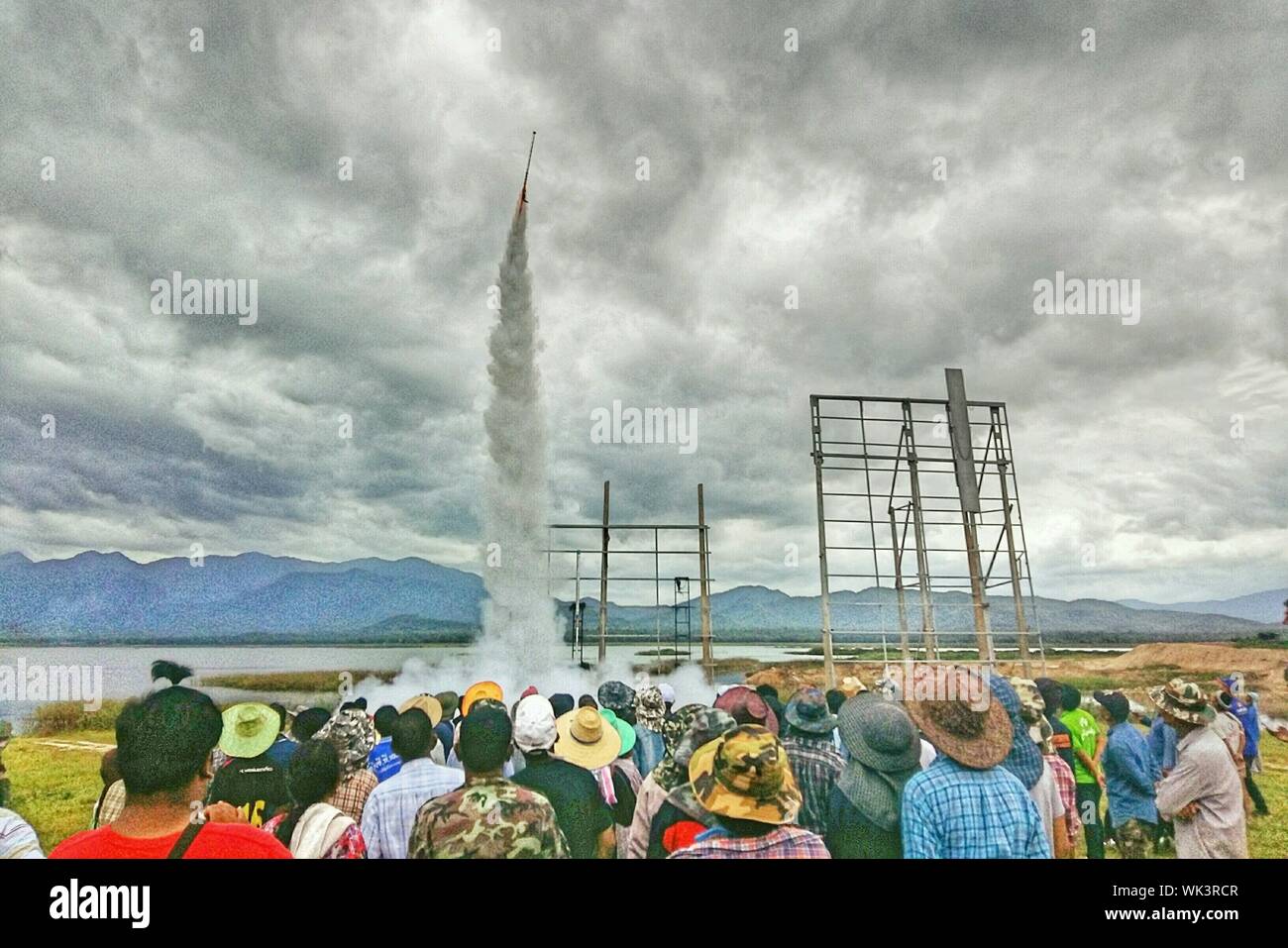 Crowd Looking At Rocket Launch At Riverbank Against Cloudy Sky Stock Photo