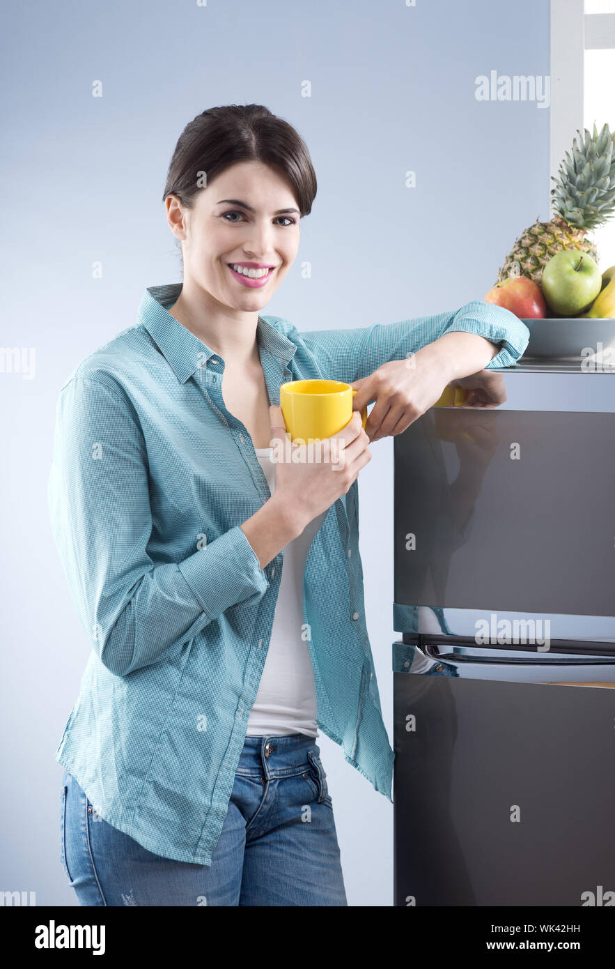 Young cheerful woman holding a yellow mug and leaning against refrigerator. Stock Photo