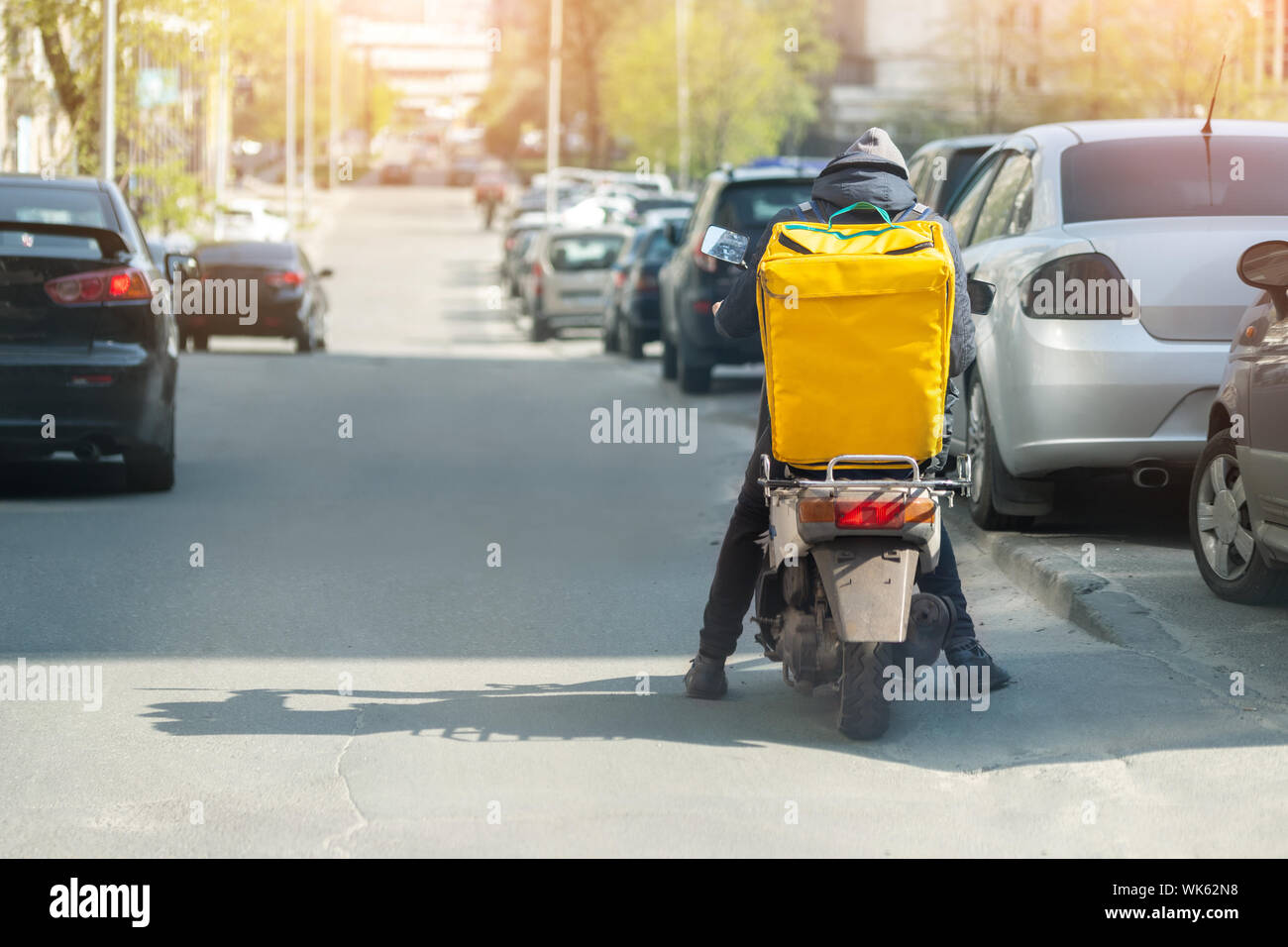 Food delivery courier with big yellow backpack riding scooter on city