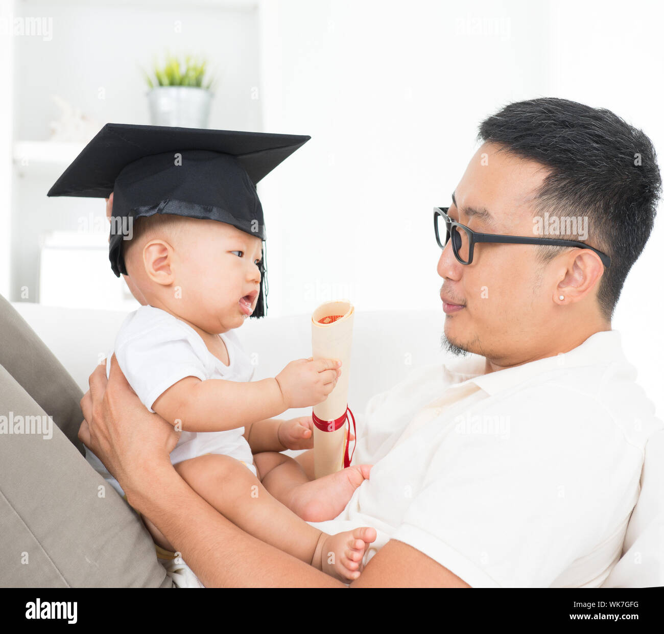 Asian family lifestyle at home. Baby with graduation cap holding certificate. Child and father early education concept. Stock Photo