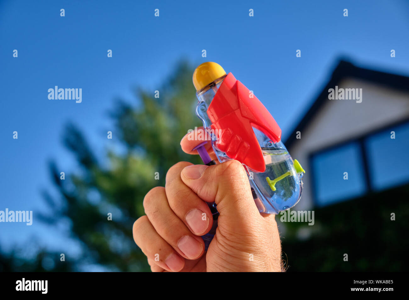 A colorful water pistol in a caucasian male hand in front of  a house and the blue sky of a sunny summer day Stock Photo