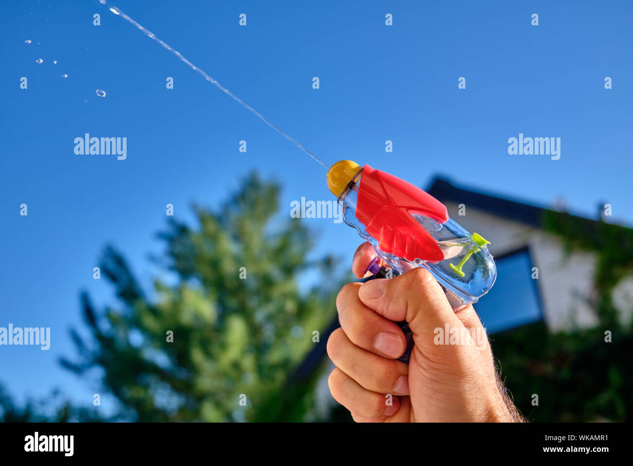 Colorful water pistol spraying out a jet of water in a caucasian male hand in front of  a house and the blue sky of a sunny summer day Stock Photo