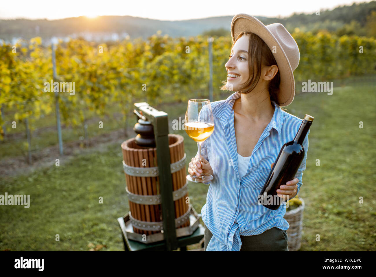 Young and cheerful woman tasting wine, while standing near the press machine on the vineyard on a sunny evening Stock Photo
