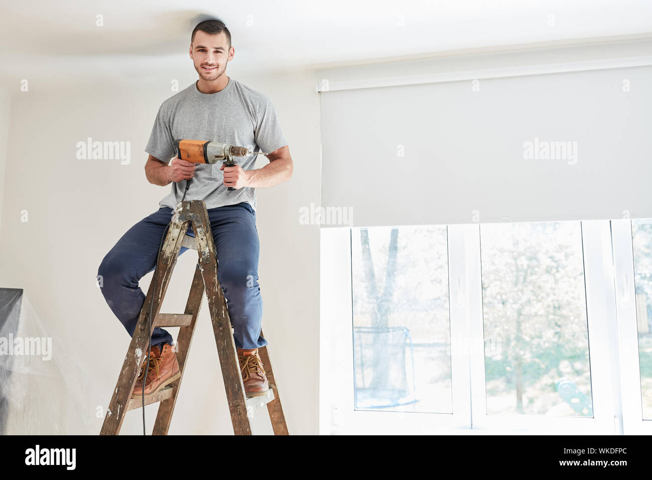 Young man as a home improvement with drill sits on a ladder at the home renovation Stock Photo