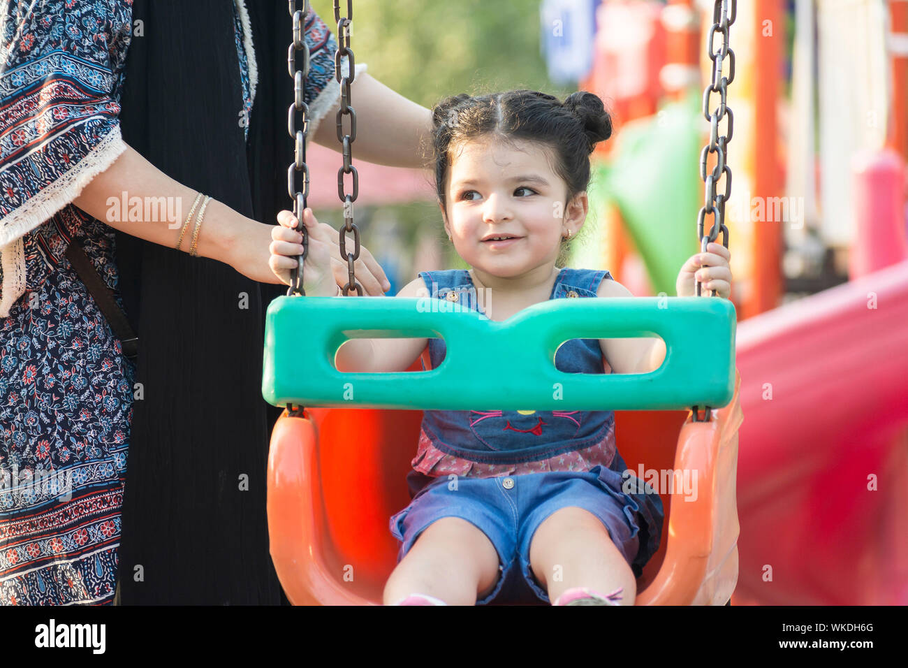 Happy little girl enjoying on swing in playground, mother support her ...