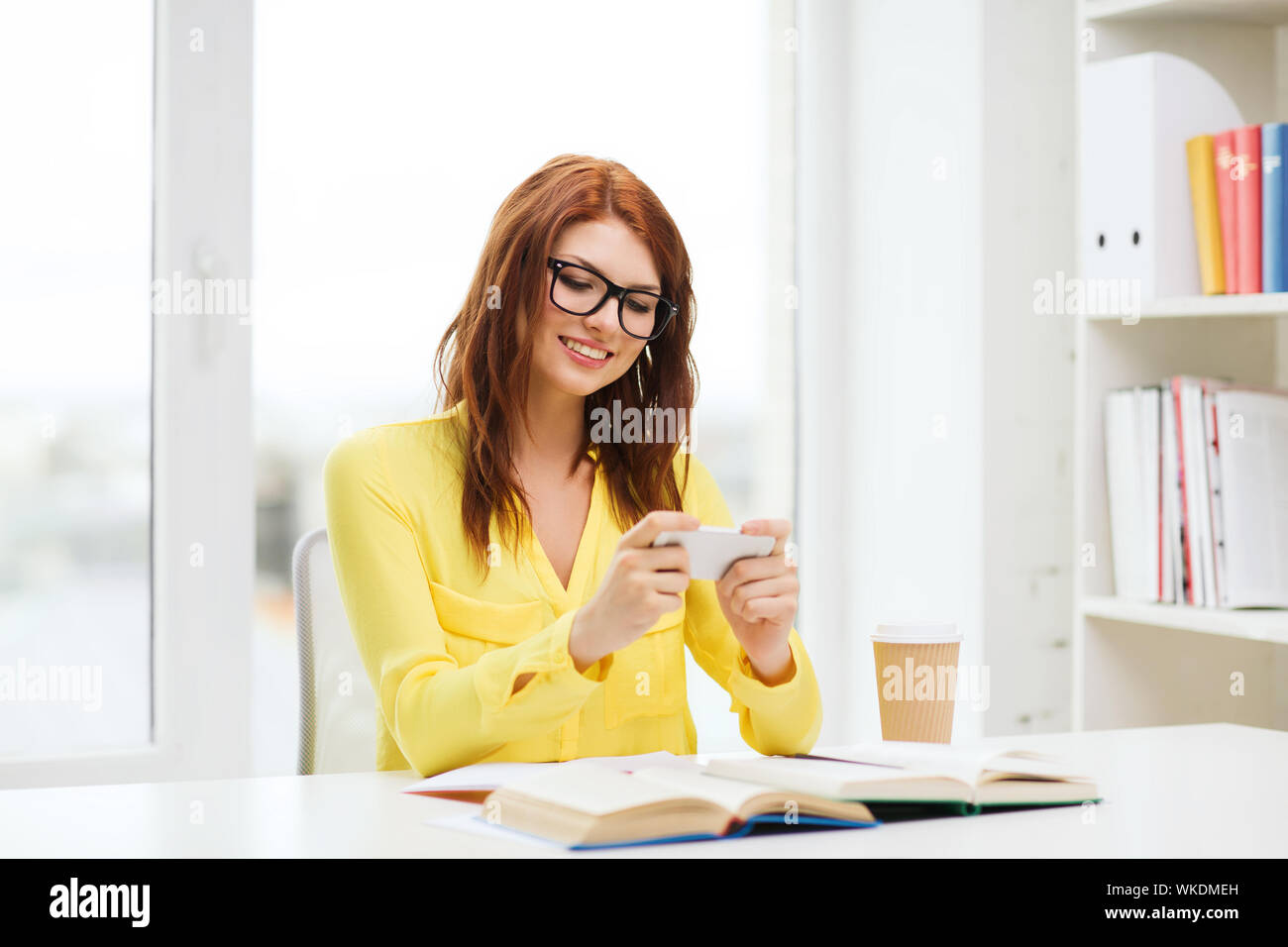 education, entretainment and technology concept - smiling student girl in eyeglasses with smartphone, books and takeaway coffee at school Stock Photo