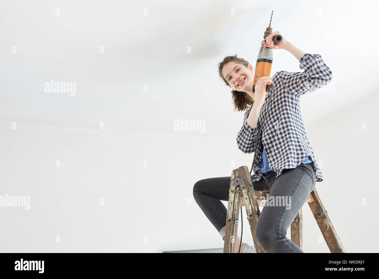 Young woman as a home improvement while drilling on the ceiling with drill sits on a ladder Stock Photo