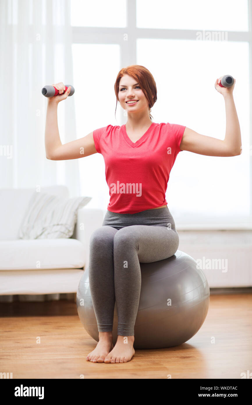 fitness, home and diet concept - smiling redhead girl exercising with fitness ball and dumbbells at home Stock Photo