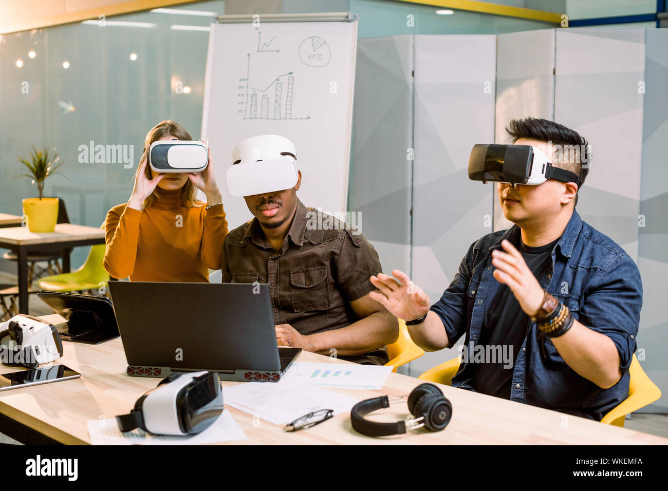 Young group of multiethnical people having fun with new technology vr headset. Caucasian girl, African and Chinese men sitting at the desk and trying Stock Photo