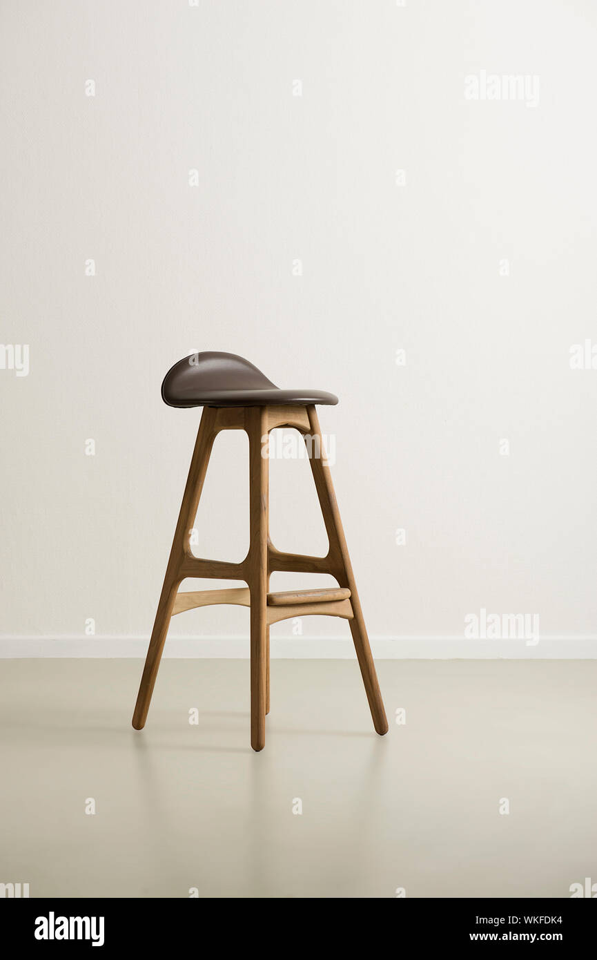 Moderrn high wooden bar stool with a black leather seat and footrests on a reflective floor in an empty room with a white wall, vertical format with c Stock Photo