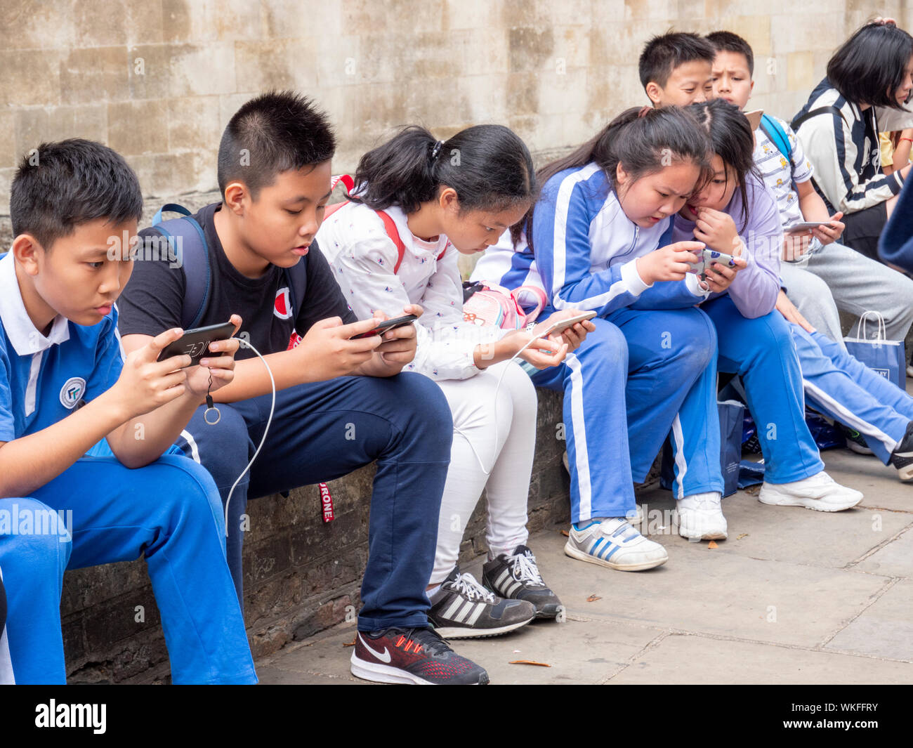 A group of young chinese tourists sitting on a wall in Cambridge UK looking at their smart phones, completely engrossed and ignoring the outside world Stock Photo