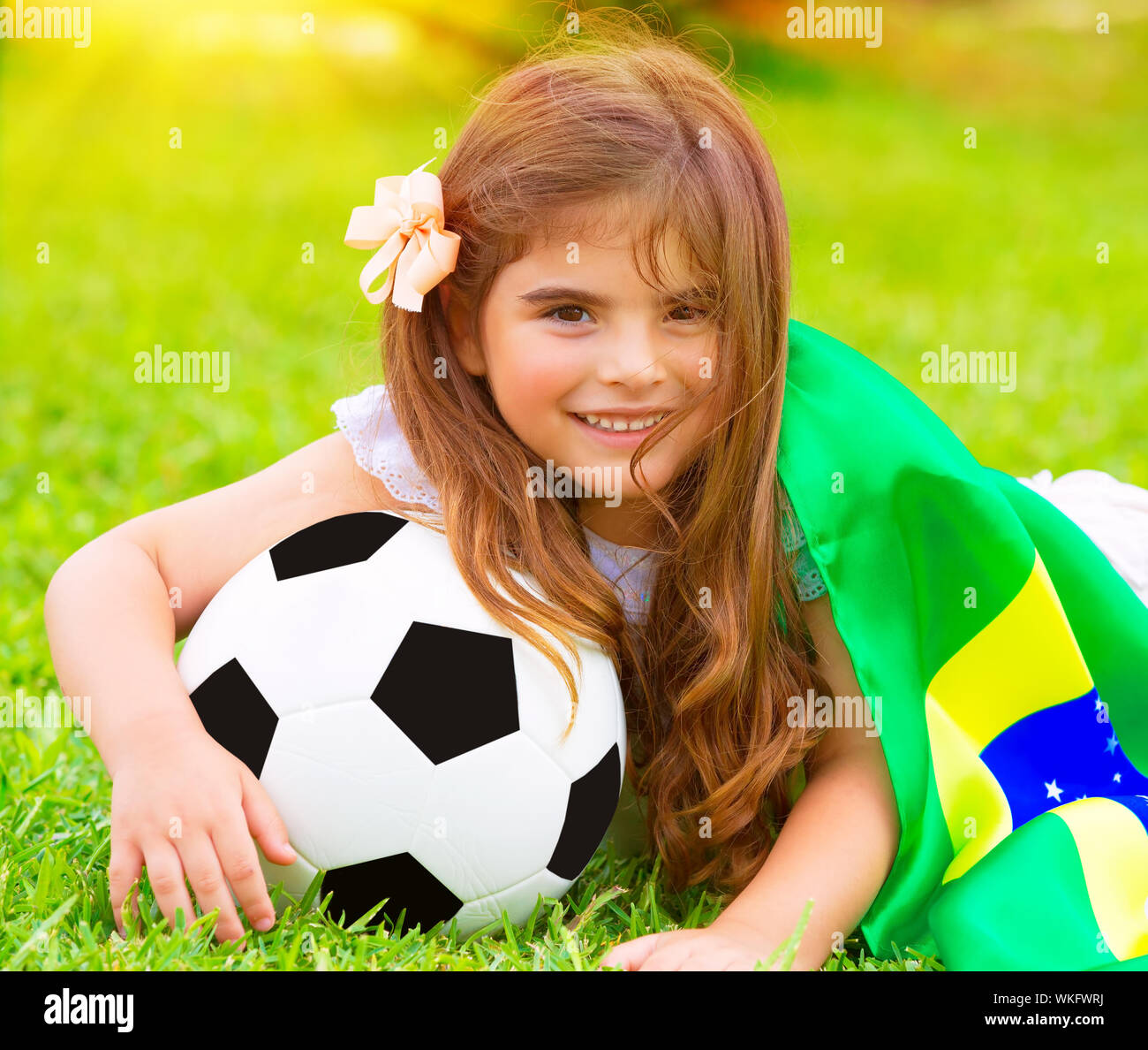 Closeup portrait of cute little football fan lying down on fresh green grass with ball and big Brazil flag, happy supporter of Brazilian football team Stock Photo