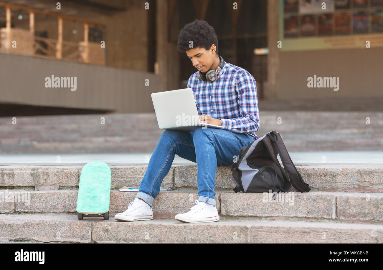 Black teen typing on laptop sitting outside near university campus Stock Photo