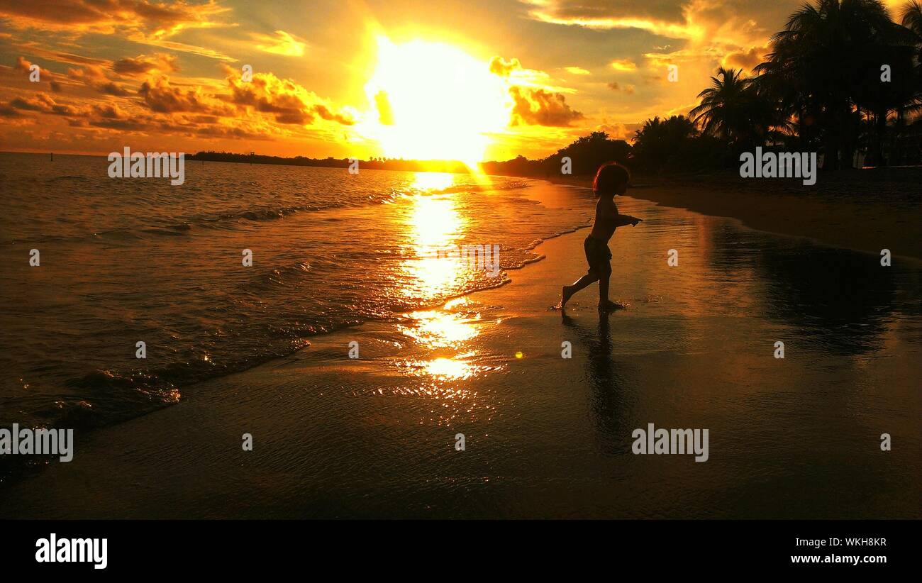 Boy Walking On Beach Against Bright Sun Sky During Sunset Stock Photo
