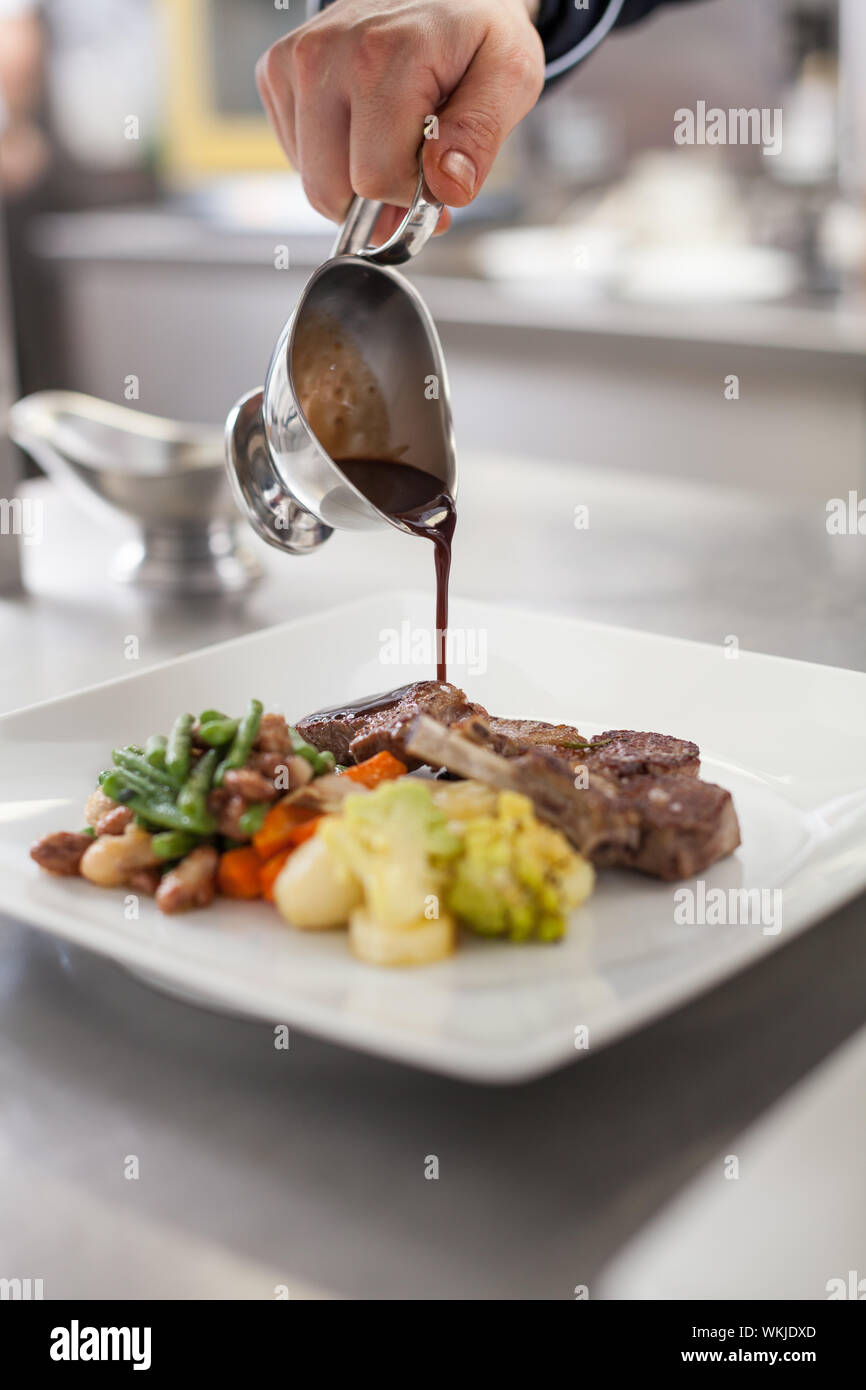 Chef plating up food in a restaurant pouring a gravy or sauce over the meat before serving it to the customer, close up view of his hand and the gravy Stock Photo