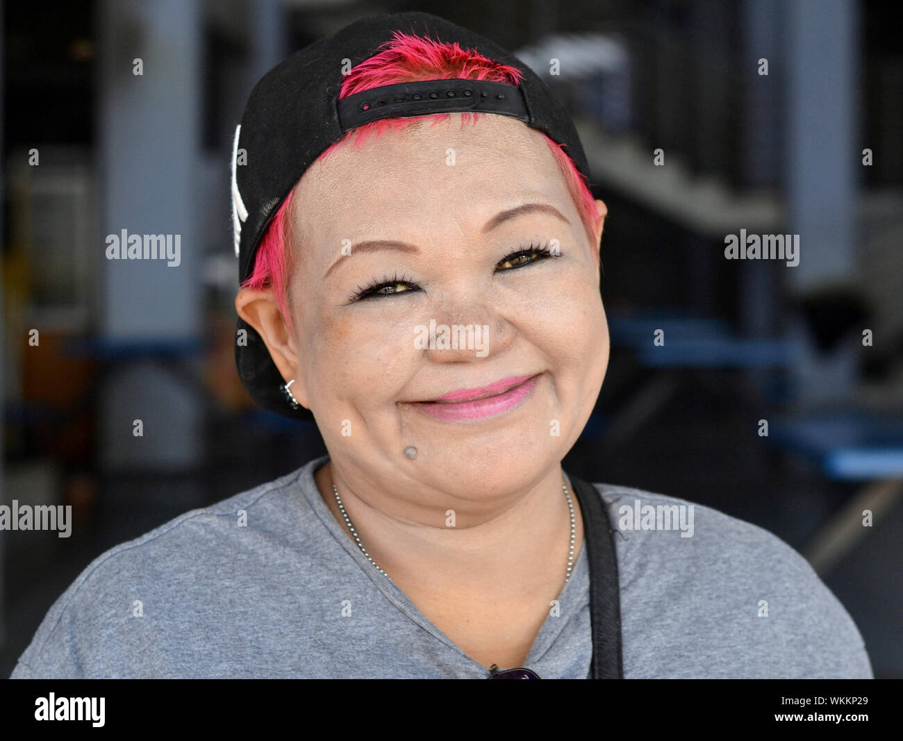 Malaysian Chinese woman with red dyed hair wears a reversed baseball cap and distinctive eye make-up and smiles for the camera. Stock Photo