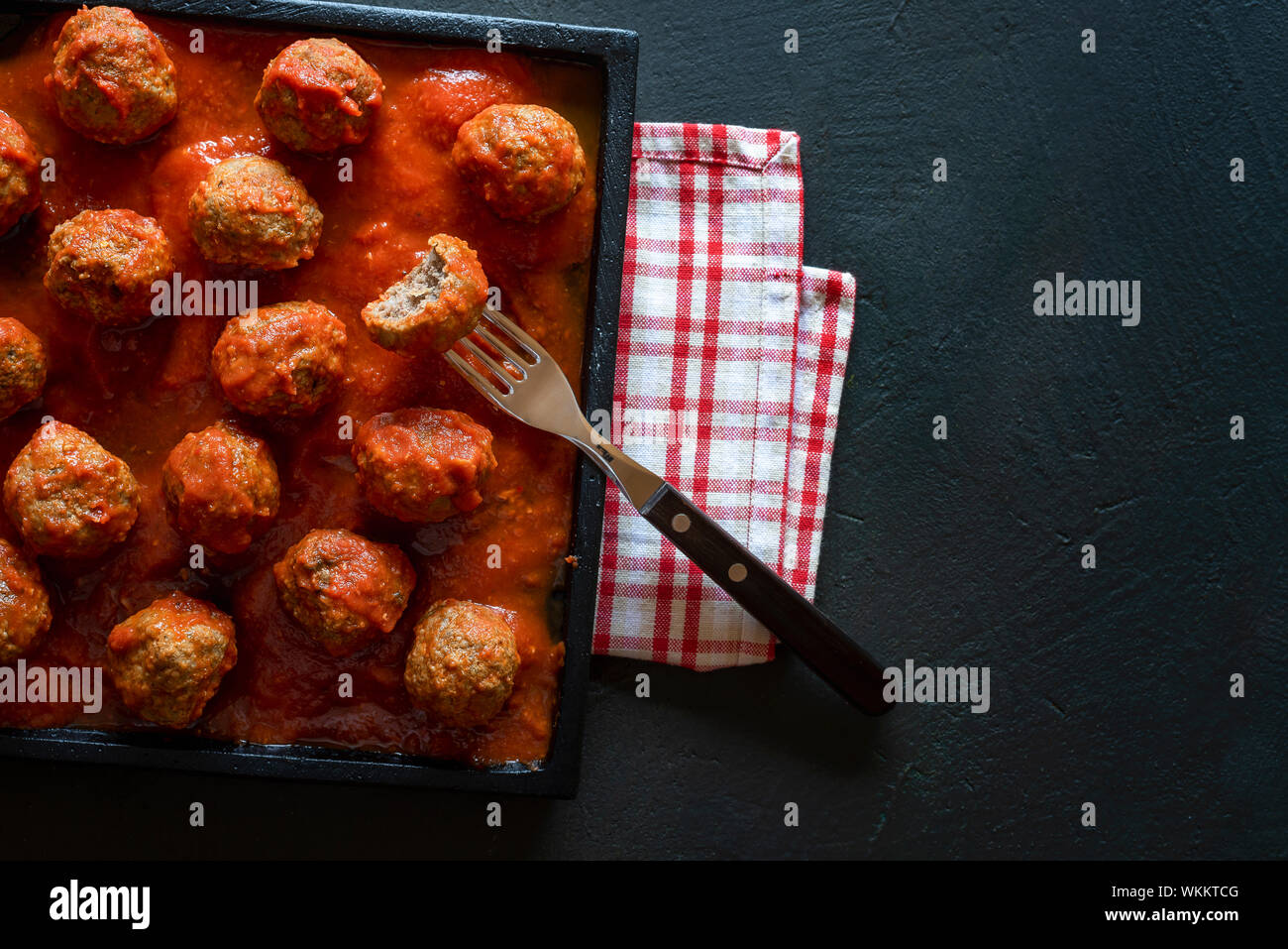 Tasty Swedish meatballs in hot spicy tomato dip on a black plate and black table. Above view of a plate of meatballs and a fork with bitten food. Stock Photo