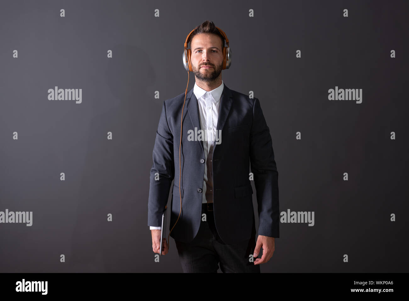A serious handsome young businessman in a suit holding a tablet and listening to music in front of a dark grey background in a studio. Stock Photo