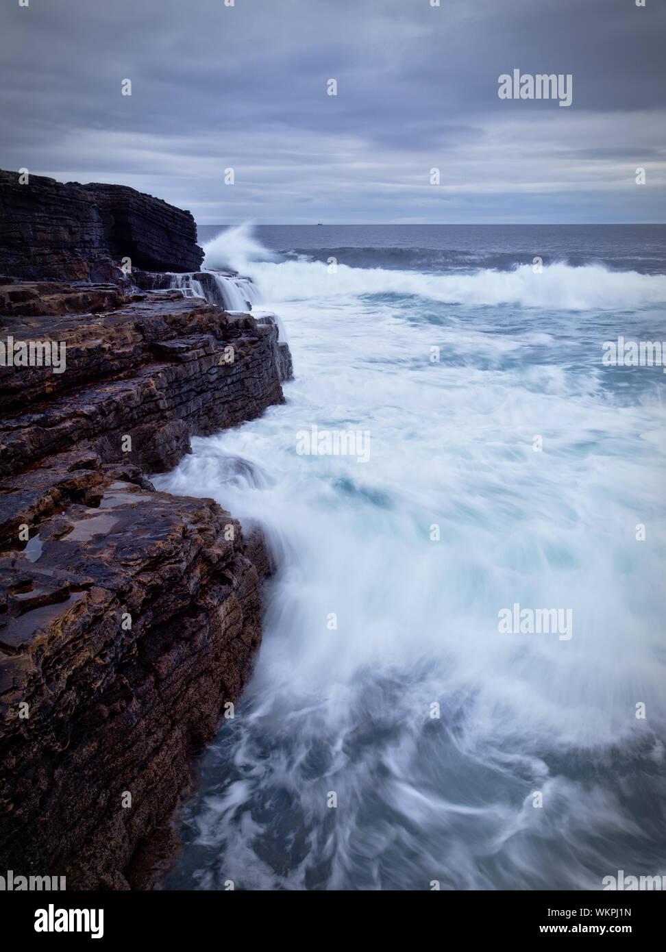 Waves crashing on the brown rocky cliffs of the Irish west coast at dusk (long exposure, portrait) Stock Photo
