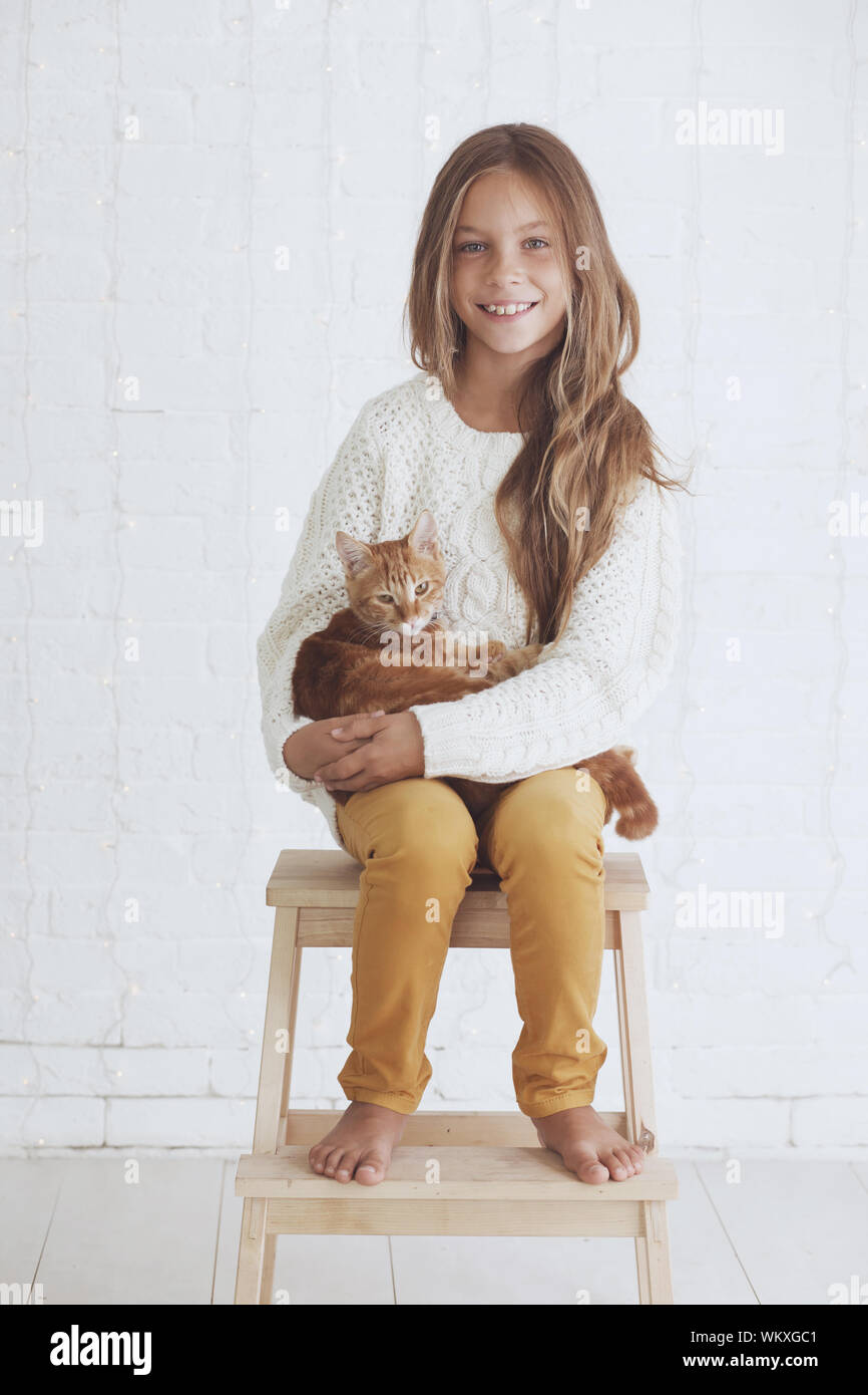 Cute teenage girl 8-9 years old wearing knit trendy winter clothes holding her ginger cat posing over white brick wall Stock Photo