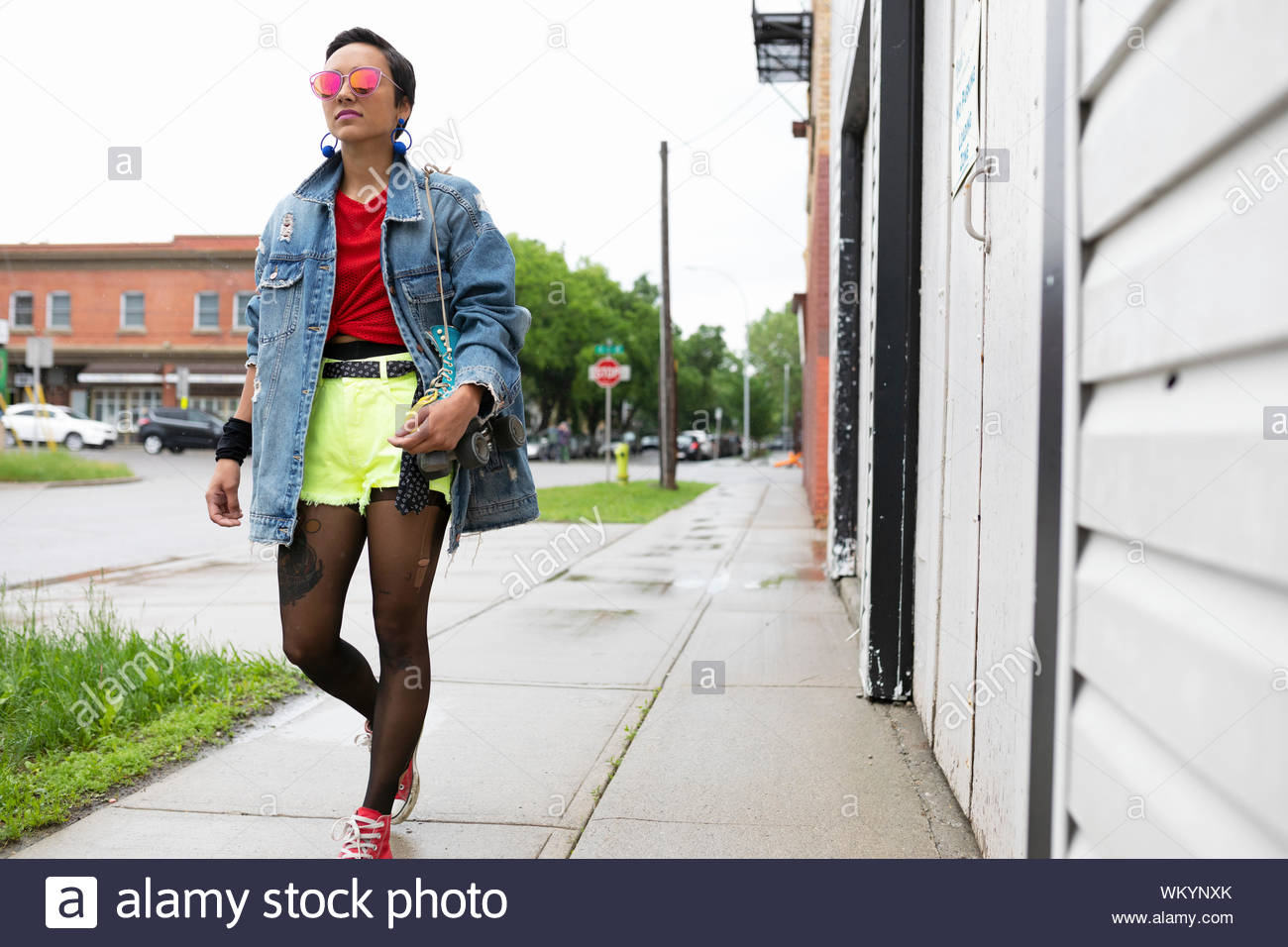 Cool young woman walking on wet sidewalk, carrying roller skates Stock