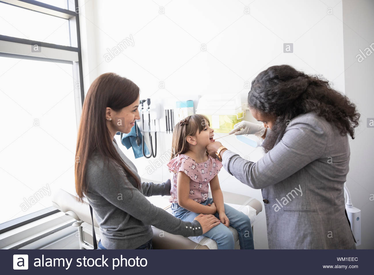 Female doctor with tongue depressor examining mouth of girl in clinic