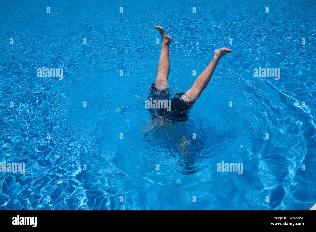 man handstands underwater with two legs peering in a blue pool Stock