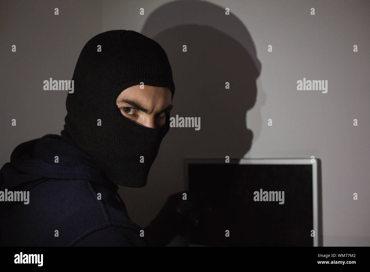Hacker in balaclava hacking laptop while looking at camera in house Stock Photo